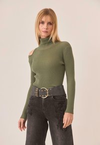 Top de cuello alto de canalé en verde oliva con un corte en un hombro. Combinado con jeans oscuros que presentan bordados florales, sujetado por un cinturón negro con tachuelas.