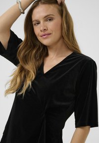 Top in velluto nero con scollatura a V profonda e maniche corte, caratterizzato da un dettaglio attorcigliato sul davanti che aggiunge texture e interesse al design.