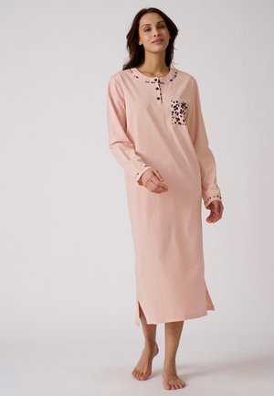 Mujer con un camisón largo rosa con cuello, puños y bolsillo en el pecho con estampados, de pie descalza frente a un fondo liso.