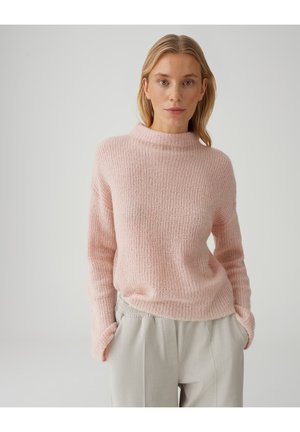 Blonde vrouw die een zachtroze gebreide trui en lichtbeige broek draagt, staand met haar handen in haar zakken tegen een effen lichte achtergrond.
