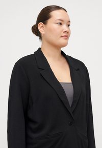 Blazer noir en tissu texturé, avec un col cranté, fermeture à un bouton et poches avant. Porté sur un haut gris.