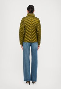 Kvinne som står med ryggen til iført en olivengrønn quiltet jakke, blå vide jeans og svarte høye hæler.