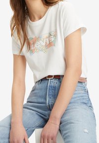 Camiseta blanca de algodón con un gráfico colorido del logo floral de Levi's. Combinada con unos jeans de mezclilla de talle alto en azul claro.