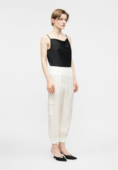 Γυναίκα όρθια που φοράει μαύρο αμάνικο τοπ, χαλαρά λινά λευκά παντελόνια με τσέπη στη μέση και μαύρες χαμηλές γόβες με μυτερή μύτη.