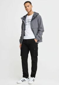 Veste légère grise avec capuche, t-shirt graphique blanc, pantalon cargo noir avec poches et baskets blanches à rayures noires.