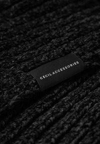 Écharpe en tricot noire avec une surface texturée. Présente une étiquette en tissu portant l'inscription "CECIL ACCESSORIES" en blanc. Le matériau semble doux et épais.