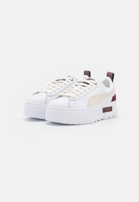 Weiße Sneakers mit einer dicken weißen Sohle, beigen Akzenten, strukturiertem Leder und brauner Markenbeschriftung an der Seite. Sie besitzen flache Schnürsenkel und eine Schlaufen-Fersenkappe.