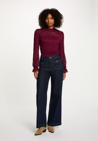 Top in pizzo bordeaux con maniche a sbuffo e scollatura a zigzag abbinato a jeans a gamba larga blu scuro con dettagli di bottoni e cuciture a contrasto.