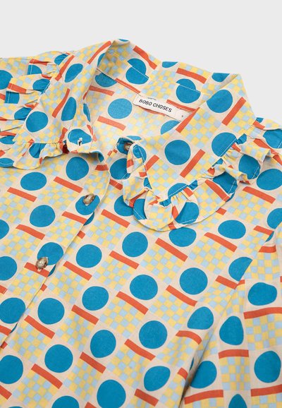 Buntes Hemd mit Knöpfen und einem Rüschenausschnitt, mit einem Muster aus blauen Kreisen und geometrischen Formen in Orange und Gelb auf einem cremefarbenen Hintergrund.