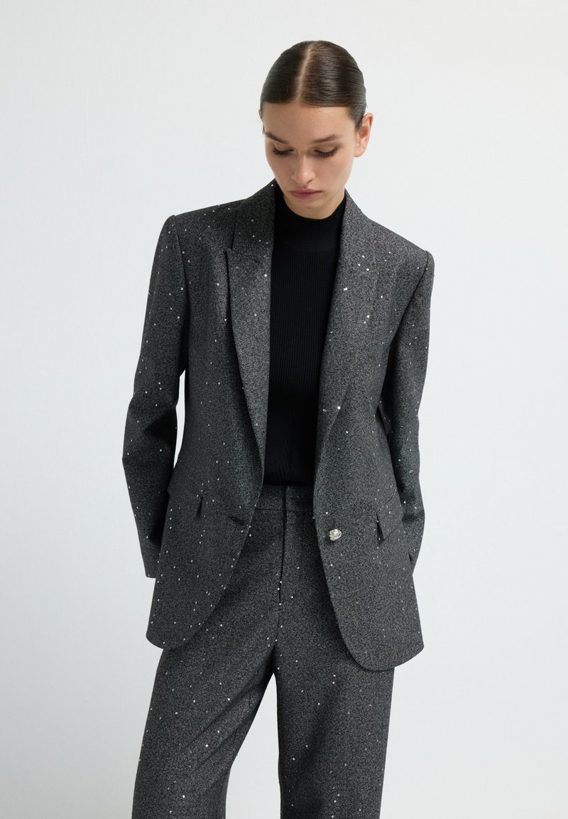 Glitzernder grauer Blazer mit taillierter Passform, versetzten Revers und metallischen Knopfdetails, kombiniert mit einem schwarzen gerippten Rollkragenpullover.
