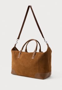 Bruine suède reistas met leren handvatten en hoeken, voorzien van een afneembare donkerbruine schouderriem, tegen een effen achtergrond.