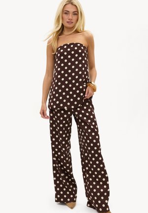Femme blonde portant un top marron sans bretelles et un pantalon large à pois blancs, des talons beiges et un grand bracelet.