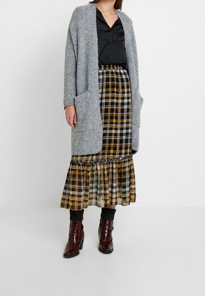 Grey knitted cardigan with pockets, paired with a yellow and black plaid midi skirt featuring ruffles. Glossy burgundy ankle boots.