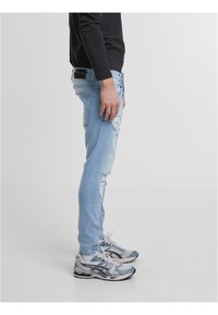 Hele sinised rebenenud teksad, vormiv lõige, kerge tuhmumisega. Kandmisel on hallid ja sinised spordijalatsid. Näha on must pika varrukaga särk.