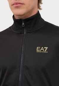 Black zip-up jacket with high collar, textured fabric, featuring a gold EA7 Emporio Armani logo on the left side.