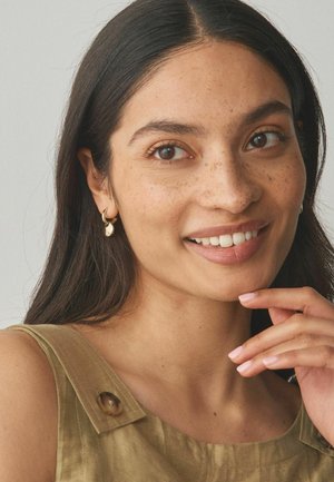 Smiling woman with long dark hair and freckles wears gold hoop earrings and an olive green sleeveless top, touching her chin gently.