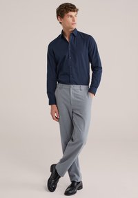 Man staat met handen in zakken, draagt een marineblauw overhemd met knopen, lichtgrijze broek en zwarte nette schoenen, tegen een effen achtergrond.