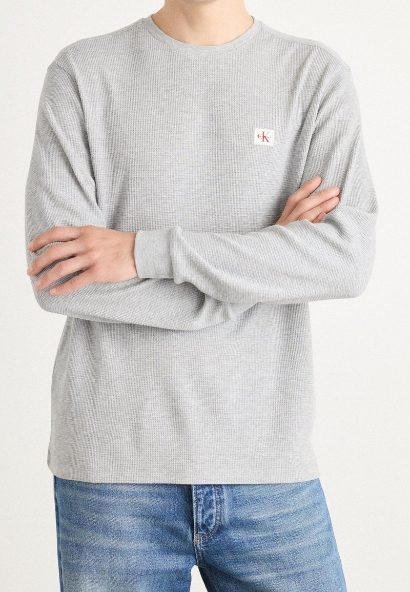 Personne aux bras croisés portant un t-shirt à manches longues côtelé gris clair avec un petit patch du logo Calvin Klein et un jean bleu.