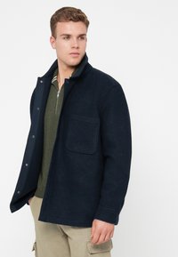 Νεαρός άνδρας με σκούρο ναυτικό μπουφάν πάνω από ελαιόχρωμο φούτερ με φερμουάρ και μπεζ παντελόνι cargo, κοιτάζοντας αριστερά σε λευκό φόντο.