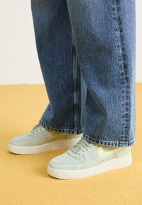 Lys grønn semsket sneakers med perforeringer og en blek gul swoosh, kombinert med vide blå denimjeans. Flatt gummisåle, teksturert stoff.