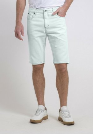 Shorts en denim vert menthe clair avec cinq poches, fermeture éclair et coutures contrastantes. Associés à des baskets blanches et une chemise blanche.