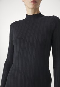 Suéter negro de manga larga con cuello ondulado, que presenta una silueta ajustada y material de punto texturizado. Diseño minimalista.