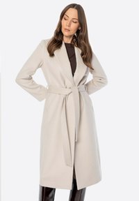 Jasno beżowy płaszcz z paskiem w talii, kołnierzem szalowym i długimi rękawami; wykonany z miękkiego materiału o gładkiej teksturze.