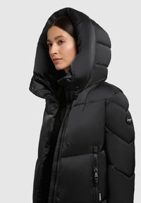 Schwarze Steppjacke mit großer Kapuze, gesteppter Textur, Frontreißverschluss und Seitentaschen. Verfügt über ein Markenzeichen auf dem Ärmel.