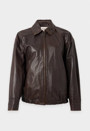 Jeune femme aux longs cheveux blonds portant une veste en cuir brun foncé, un haut vert, une jupe midi fleurie et des bottes noires, debout sur un fond blanc.