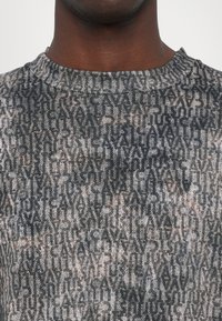 Siv sweater z značilnim teksturiranim materialom z ponavljajočim se besedilnim vzorcem v temnejših odtenkih. Oblika s šobastim vrhom in subtilnim, sproščenim krojem.