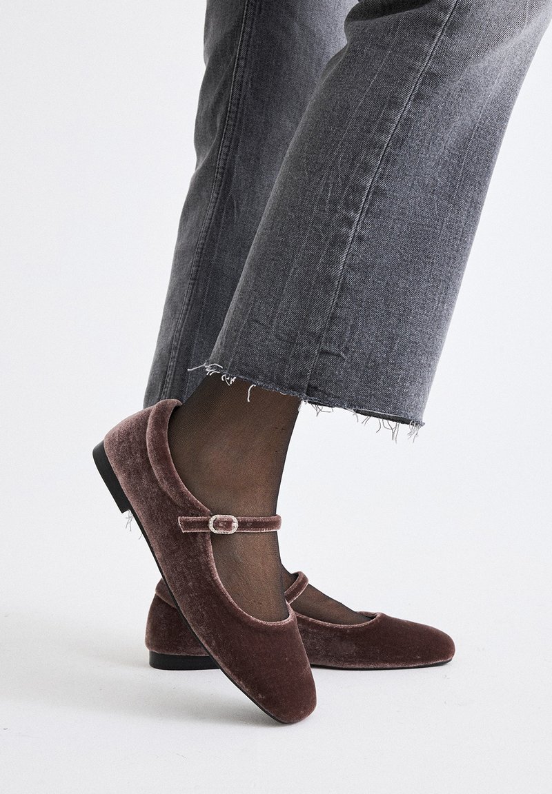 Zapatos planos estilo Mary Jane en terciopelo burdeos, con punta redondeada, correa de tobillo con hebilla y suela baja negra. Combinados con jeans grises deshilachados.