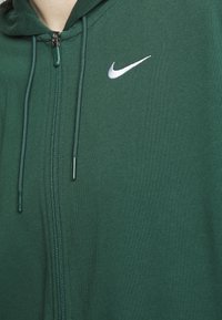 Haut à fermeture éclair vert en tissu doux. Présente un logo Nike blanc et des cordons de serrage. A une texture lisse avec un design ajusté.