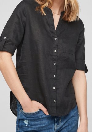 Femme portant une chemise noire boutonnée avec manches retroussées et un jean bleu déchiré, une main négligemment dans la poche.