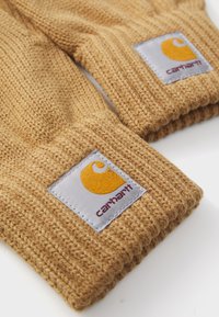 Pruunid kudumisega kindad, millel on soonikkoest tekstuur, hall kangast silt, millel on kollane logo ja burgundi värvi "Carhartt".