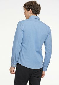 Vaaleansininen farkkupaita pitkillä hihoilla, napilliset hihansuut, kaulus ja kaareva helma. Liukas pinta, jossa on hienovaraisia ompelutarkkuuksia.