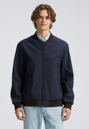 Junger Mann mit lockigem Haar, der eine marineblaue Bomberjacke über einem gestreiften Hemd und hellblauen Jeans trägt und vor einem schlichten Hintergrund steht.