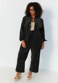 Zwarte corduroy oversized shirt en bijpassende wijde broek, textuurstof, twee borstzakken, gecombineerd met lichtbeige platform schoenen.