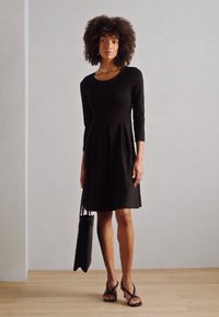 Black long-sleeve dress with a fitted bodice and flared skirt. Smooth fabric, paired with black sandals and a structured black tote.