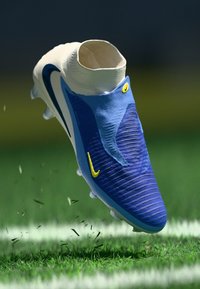 Chuteira de futebol azul e branca com design de tornozelo alto, apresentando material texturizado, acentos amarelos e forma aerodinâmica, pairando acima da relva.