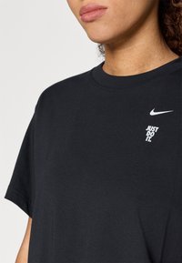 Μαύρη βαμβακερή μπλούζα με κοντό μανίκι και στρογγυλή λαιμόκοψη, με λευκό λογότυπο Nike και την επιγραφή "JUST DO IT" σε έντονη, σύγχρονη γραμματοσειρά.