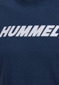Marinblå t-shirt i bomull med kraftig vit text "HUMMEL" i en sportig, versal stil över bröstet. Mjuk textur, rund halsdesign.