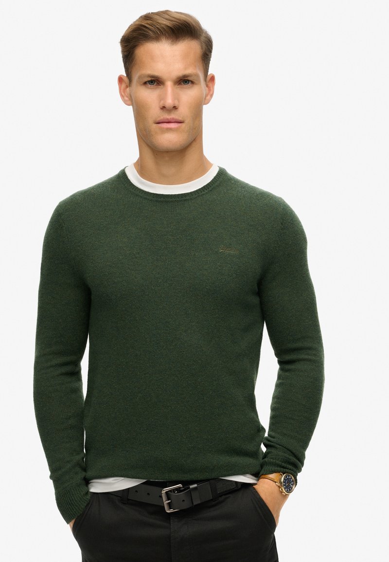 Groene ronde hals trui met een tekstuur gebreide patroon, gedragen over een wit shirt. Kenmerkt ribbels aan de manchetten en een subtiele logo op de borst.