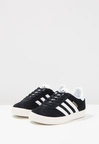 Zapatillas de ante negro con tres franjas blancas, suela de goma texturizada y texto dorado "Gazelle" en el lateral. Detalles en blanco en la lengüeta.