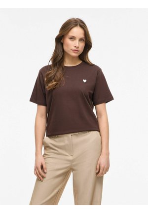 Donna con lunghi capelli castani che indossa una t-shirt marrone scuro con un piccolo cuore bianco sul petto e pantaloni beige, in piedi davanti a uno sfondo bianco.