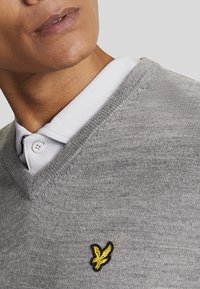 Maglione grigio lavorato a maglia con scollatura a V, caratterizzato da una sottile texture e un logo di aquila ricamato di colore giallo sul lato sinistro del petto. Collo bianco sotto.