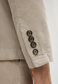 Beżowy mankiet marynarki ze sztruksu z czterema ciemnymi guzikami, teksturowaną tkaniną i delikatnym wzorem prążków o dopasowanym kroju.