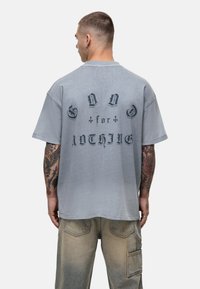 Szara, oversize koszulka z krótkim rękawem i okrągłym dekoltem, z czarnym nadrukiem na plecach, wykorzystującym różne czcionki. Casualowy styl.