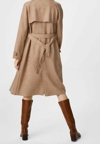 Beżowy zamszowy płaszcz trench z paskiem w talii, długimi rękawami i warstwowym pom designem z tyłu. Brązowe kozaki do kolan uzupełniają stylizację.
