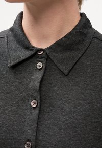 Gros plan d'une personne portant une chemise à col gris foncé avec quatre boutons visibles fermés à l'avant.