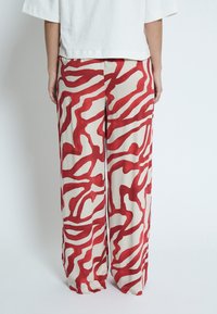 Rote und beige gemusterte Hosen mit einem fließenden, weiten Beinanschnitt, die abstrakte wellenförmige Linien zeigen. Das Material wirkt leicht und glatt.